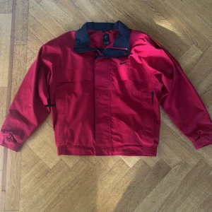 Vintage Nike vindjacka. - Riktigt snygg röd vindjacka från Nike med svart krage och diskret svart swoosh-logga på bröstet. Jackan har dragkedja och tryckknappar framtill, elastisk midja och ärmslut samt praktiska fickor. Perfekt för dig som gillar sportig och clean stil.