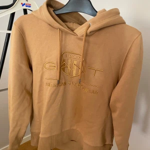 Beige hoodie från GANT - Säljer en snygg beige hoodie från GANT med broderad logga och text framtill. Tröjan har huva med snörning och ribbade muddar. Perfekt för chill dagar eller när du vill ha en clean och stilren look.