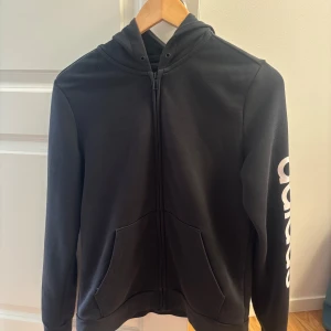 Svart hoodie från Adidas med tryck - Svart hoodie från Adidas med vit logga längs ena ärmen. Tröjan har dragkedja framtill, huva och två fickor. Tillverkad i mjuk bomullsblandning, perfekt för chill eller träning. Klassisk och stilren design med normal passform.