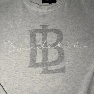 Ljusgrå stickad tröja från Bondelid - Ljusgrå stickad tröja från Bondelid med stort broderat logotyptryck och text på bröstet. Tröjan har rund halsringning, ribbade muddar och lång ärm. Perfekt för dig som gillar klassisk och stilren design.