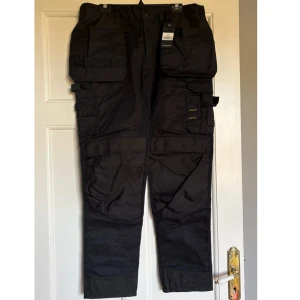 Svarta arbetsbyxor - Byxor i storlek 54, helt nya med lapp kvar eftersom dom inte passade. Beskrivning: Arbetsbyxa med många fickor och fästpunkter. Den här byxan från Bluewear är av en kraftig, slittålig och stretchig modell som dessutom är OEKO-TEX-certifierad. För att du ska kunna ha allt du behöver nära till hands har vi försett byxan med många smarta fickor och fästpunkter!