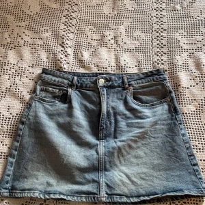 Blå jeanskjol från H&M - Snygg blå jeanskjol från H&M med klassisk ficksdesign och knappstängning framtill. Kjolen har en lätt A-linjeform och bälteshällor i midjan. Perfekt för dig som gillar en enkel och tidlös stil.