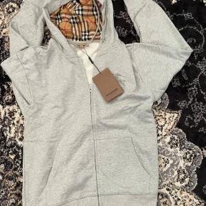 Burberry hoodie - Grå hoodie från Burberry med klassiskt rutigt foder i huvan och broderad logga på bröstet. Tröjan har hel dragkedja, snörning i huvan och kängurufickor framtill. Perfekt för en chill och stilren look. Helt ny oanvänd 