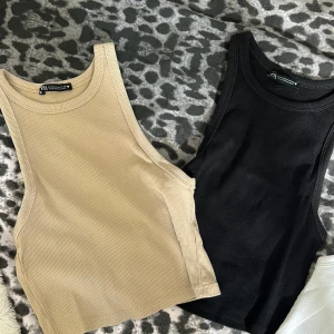 Ribbade linne-toppar från Zara - Två ribbade linne-toppar från Zara, en i beige och en i svart. Båda är croppade med hög halsringning och ärmlösa. Perfekta att styla med jeans eller kjol för en clean look. Säljes tillsammans eller separat.