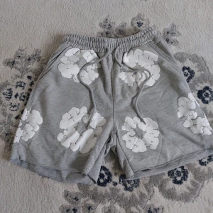 Grå shorts med blommor från Denim Tears - Grå mjuka shorts från Denim Tears med vita blommiga tryck över hela plagget. De har elastisk midja med snörning och en bakficka. Perfekta för chill dagar och har en avslappnad passform.