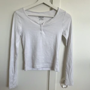 Vit långärmad topp från H&M - Säljer en enkel och stilren vit långärmad topp från H&M. Toppen har en rundad halsringning med knappdetalj framtill och är tillverkad i mjuk bomull. Perfekt att matcha med jeans eller kjol för en clean look.
