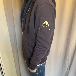 Äkta! Mörkblå hoodie från Moose Knuckles - Säljer en mörkblå hoodie från Moose Knuckles med dragkedja, huva och mjukt foder. Tröjan har två stora fickor fram och en snygg logga i metall på ärmen. Perfekt för kyliga dagar såsom vintern samt hösten. Scan funkar och den är i mycket bra skick! Hör gärna av er vid frågor!🙌🙌