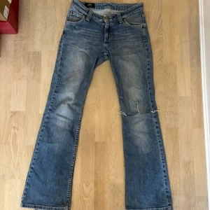 Blå bootcut jeans från Lee - Snygga blå jeans från Lee med bootcut passform och klassisk femficksdesign. Jeansen har slitningar och en trasig detalj vid knät för en cool, avslappnad look. Tillverkade i bomull med Lee-logga på bakfickan och läderpatch i midjan. Storlek 36!🩷