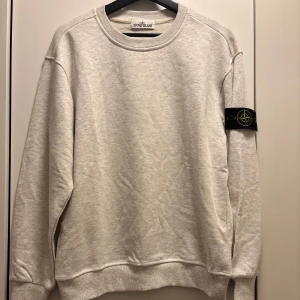 Stone Island Grå sweatshirt  - Snygg ljusgrå sweatshirt från Stone Island med klassisk rund hals och ikonisk logga på vänster ärm. Tröjan har ribbade muddar och är tillverkad i mjuk bomull för maximal komfort. Perfekt för dig som gillar stilrena och trendiga plagg.