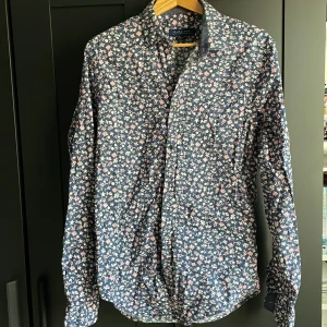 Blommig skjorta från Zara Man - Snygg långärmad skjorta från Zara Man med blå botten och blommigt mönster i vitt och rosa. Skjortan har klassisk krage, knäppning framtill och är tillverkad i mjuk bomull. Perfekt för dig som vill sticka ut med en cool och färgglad look.