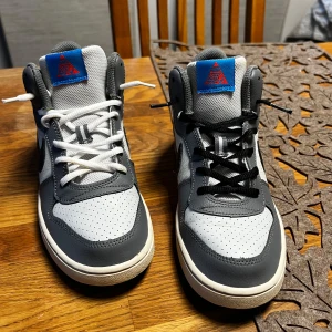 Nike grå och vita sneakers strl 37.5 - Nike sneakers i grått och vitt med svart Swoosh och blå/röd detalj på plösen. Skorna har perforerad tå, snörning (ena skon med vita och andra med svarta skosnören) och klassisk rund tå. Ovandel i syntet och mesh för bra andningsförmåga. Perfekt för dig som gillar streetstyle.