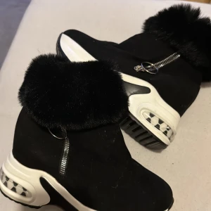 Svarta boots med fuskpäls och dragkedja - Säljer ett par svarta boots med chunky vit sula och coolt rutmönster i sulan. Skorna har mjuk fuskpäls runt skaftet och dragkedja med metallring på sidan. Perfekta för dig som vill sticka ut med en sportig men ändå trendig look.