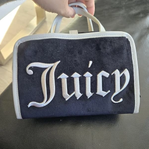 Juicy Couture svart sammetshandväska - Snygg svart handväska från Juicy Couture i mjuk sammet med vita detaljer och stort broderat Juicy-tryck på framsidan. Väskan har dubbla handtag, dragkedja med JC-berlock och insida med logomönster. Perfekt för dig som gillar en ikonisk Y2K-stil.
