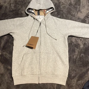 Grå hoodie från Burberry med dragkedja - Snygg ljusgrå hoodie från Burberry med klassiskt rutigt foder i huvan och broderad logga på bröstet. Tröjan har hel dragkedja, snörning i huvan och två fickor framtill. Perfekt för dig som vill ha en stilren och exklusiv look.