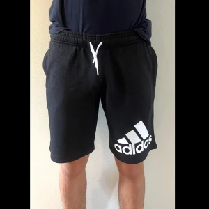 Svarta Adidas shorts med snörning - Svarta shorts från Adidas med vit logga på ena benet och vit snörning i midjan. Modellen har elastisk midja och två sidofickor. Perfekta för träning eller chill. Mjukt bomullsmaterial som känns skönt mot huden.