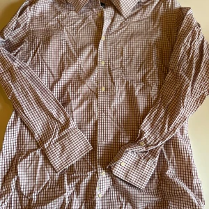 Rutig skjorta från Lapidus, stl 44 - Klassisk vit skjorta från Lapidus med vinröda rutor och knappar framtill. Skjortan har lång ärm, bröstficka och krage. Tillverkad i bomull och är non-iron, vilket gör den enkel att sköta. Perfekt för dig som gillar stilrena och tidlösa plagg.