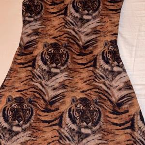 Brun klänning med tigerprint - Unik klänning med all-over tigerprint i bruna och svarta toner. Klänningen har en figurnära passform och är tillverkad i ett mjukt material som känns skönt mot huden. Perfekt för dig som vill sticka ut med ett djurmönster och cool attityd.