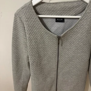 Grå cardigan jacka från Vila - Säljer en ljusgrå cardigan jacka från Vila med snyggt strukturerat tyg och dragkedja framtill. Jackan har rund halsringning och långa ärmar, perfekt för lager på lager. Enkel och stilren design som passar till det mesta.