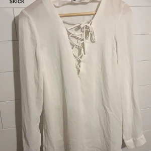 Vit långärmad skjorta med snörning - Vit långärmad skjorta med snygg snörning framtill i v-ringad modell. Skjortan har en clean och stilren look, perfekt för dig som gillar minimalistisk stil. Materialet känns lätt och luftigt, vilket gör den skön att bära.