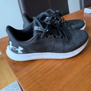 Svarta sneakers träningsskor från Under Armour - Svarta sneakers från Under Armour med vit sula och tydlig logga på hälen. Ovandelen är i textil med diskret randig struktur och snörning framtill. Skorna har en sportig look och är lätta med komfortfoder, perfekta för träning eller vardag.