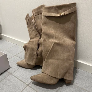 Beige mockastövlar med klack - Helt oanvända, med paketet. Snygga beige stövlar i mocka med spetsig tå och bred klack. Stövlarna har en dekorativ guldfärgad detalj upptill och dragkedja på insidan. Perfekta för att lyfta din outfit med en trendig touch. 
