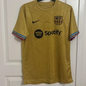 FC Barcelona Lewandowski guldtröja Nike - Snygg FC Barcelona fotbollströja i guld med svarta detaljer, Lewandowski 9 på ryggen och Spotify-logga på bröstet. Tröjan har korta ärmar med färgglada ränder vid muddarna och är tillverkad i ett lätt, glansigt material. Perfekt för dig som älskar Barca och vill sticka ut.