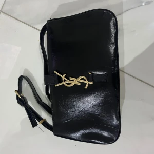 Svart axelväska - Snygg svart. Väskan har justerbar axelrem med dekorativ tofs och stilren, rektangulär form. Perfekt för dig som vill ha en klassisk och trendig accessoar.