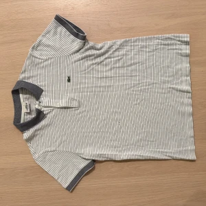 Randig pikétröja från Lacoste - Snygg vit och marinblå randig pikétröja från Lacoste med klassisk krage och knäppning framtill. Tröjan har korta ärmar och den ikoniska gröna krokodilloggan broderad på bröstet. Regular fit och mjukt bomullsmaterial. Det finns små fläckar på magen. Se bild.