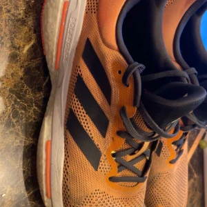 Adidas Solar Glide orange sneakers - Säljer ett par Adidas Solar Glide sneakers i orange mesh med svarta detaljer och klassiska tre ränder på sidorna. Skorna har svart snörning, vit sula med orange inslag och svart plös med gul Adidas-logga. Perfekta för löpning eller vardagsstil. Använda 2 gånger . Nypris runt 2000