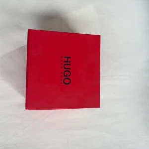 Hugo Boss klocka med brun läderrem - Stilren klocka från Hugo Boss med svart urtavla och silverfärgat hölje. Klockan har två mindre urtavlor och en brun läderrem med struktur. Levereras i original röd box med Hugo Boss-logga. Perfekt accessoar för en clean och modern look.