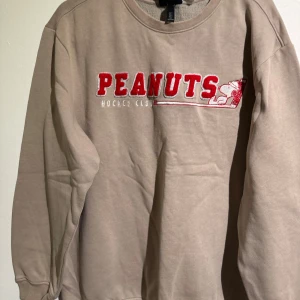 Beige Peanuts sweatshirt med tryck - Beige sweatshirt med rött och vitt Peanuts-tryck fram och Van Pelt's Proshop-tryck bak. Rund halsringning, långa ärmar och mjukt bomullsmaterial. Perfekt för dig som gillar Snoopy och vill ha en chill och snygg tröja med retrovibbar.