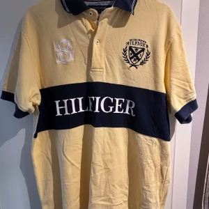 Gul och marinblå Hilfiger piké - Säljer en gul och marinblå t-shirt från Tommy Hilfiger i storlek M. T-shirten har korta ärmar, klassisk krage med knappar och ett stort Hilfiger-tryck över bröstet. Broderad logga och emblem på framsidan. Tillverkad i mjuk bomull.
