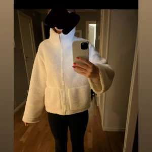 Vit teddyjacka med dragkedja - Mysig vit teddyjacka från lager 157 med hög krage och två stora fickor framtill. Jackan har rak passform, lång ärm och stängs med dragkedja. Perfekt för kyliga dagar när du vill vara både varm och snygg.
