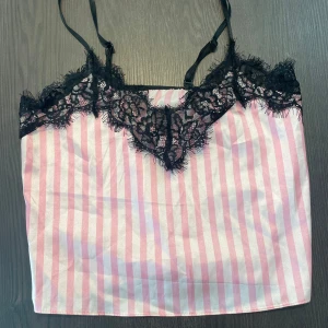Rosa randig linnetopp med spets - Söt linnetopp med rosa och vita ränder, tunna axelband och svart spetsdetalj längs urringningen. Toppen har en lätt glansig finish och justerbara band för perfekt passform. Perfekt för en trendig och feminin look.