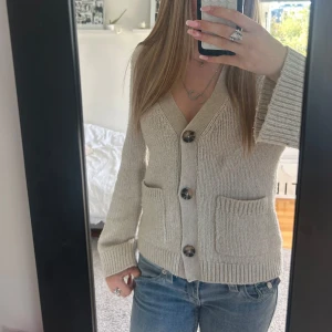 Beige stickad kofta med knappar - Supermysig beige stickad kofta med stora knappar framtill och två fickor. Koftan har v-ringning och långa ärmar, perfekt att slänga på sig över en t-shirt eller linne. Enkel och stilren design som passar till allt.