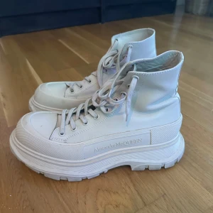 Vita höga sneakers Alexander McQueen - Säljer ett par vita höga sneakers från Alexander McQueen med chunky platt sula och snörning. Skorna är i skinn och har rund tå samt tydlig logga på sidan och tungan. Perfekta för dig som gillar statement-sneakers med modern vibe.