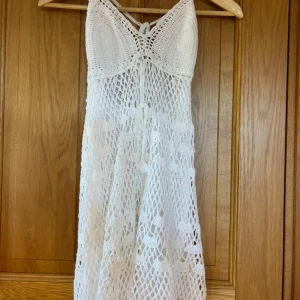 Vit virkad kortklänning med knyt - Supersöt vit virkad klänning med snyggt nätmönster och knyt i ryggen. Klänningen är ärmlös och har en lätt och luftig känsla, perfekt för varma dagar. Den har en feminin passform och fina detaljer i virkningen som gör den extra unik. Köpt second hand ingen storleks lapp men skulle uppskatta den är i storlek S/M