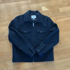 Svart jacka från River Island, M - Stilren svart jacka från River Island med klassisk krage, två bröstfickor med knappar och dragkedja framtill. Jackan har raka linjer och lång ärm, perfekt för dig som gillar en clean look. Materialet är slätt och känns bekvämt.