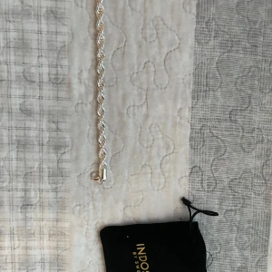 Silverfärgat armband från Indossa - Snyggt armband i silverfärg från Indossa by Sweden med tvinnad kedja och robust lås. Kommer med svart tygpåse med guldfärgad logga. Perfekt accessoar för dig som gillar stilrena detaljer och vill ha något som sticker ut lite extra. 21 cm långt, 8 mm. 