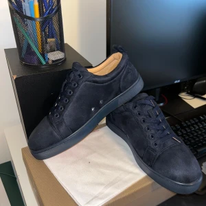 Mörkblå mocka sneakers Louboutin - Snygga mörkblå sneakers från Christian Louboutin i mjuk mocka med klassisk röd sula. Skorna har rund tå, snörning och diskret broderad logga på hälen. Insidan är beige och sulan är i gummi. Perfekta för dig som vill ha en lyxig och stilren look.