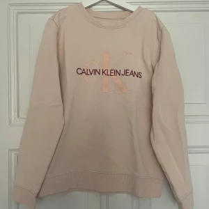 Tröja från Calvin Klein Jeans - Säljer en beige sweatshirt från Calvin Klein Jeans med stor logga framtill i ljusrosa och röd text. Tröjan har rund halsringning, långa ärmar och ribbade muddar. Perfekt för en chill och stilren look. Nypris: 800, utgångspris 400. Den har använts några gånger men den ser oanvänd ut. Säljer då den är för liten 💗
