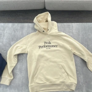 Beige hoodie från Peak Performance - PASSAR XS VÄLDIGT BRA 😊………..        Snygg beige hoodie från Peak Performance med broderad logga på bröstet. Tröjan har huva med dragsko och en stor magficka. Perfekt för dig som gillar en clean och stilren look. Mjuk insida och ribbade muddar för extra komfort.