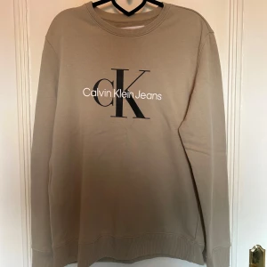 Beige Calvin Klein sweatshirt - Snygg beige sweatshirt från Calvin Klein Jeans med stor svart CK-logga och vit text på bröstet. Storlek L. 