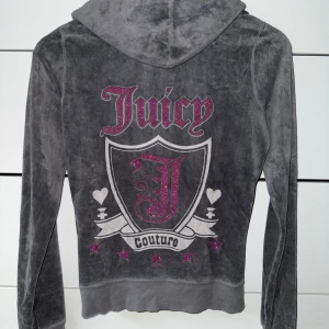 vintage Juicy Couture grå velour hoodie - Juicy Couture hoodie i mjuk grå velour med glittriga rosa detaljer och stort tryck på ryggen. Tröjan har huva, dragkedja framtill och fickor. Perfekt för dig som vill ha en ikonisk och bekväm look med lite extra bling.