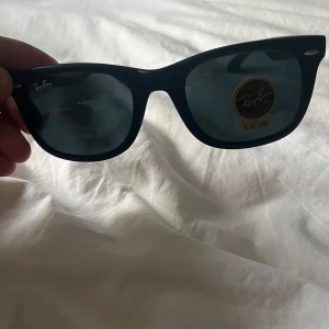 Svarta Ray-Ban Wayfarer solglasögon - Klassiska svarta Ray-Ban Wayfarer solglasögon med mörka glas och ikonisk form. Modellen är RB2140 och tillverkad i Italien. Snygg och tidlös design som passar till många stilar. Ray-Ban-logga på både lins och skalmar.