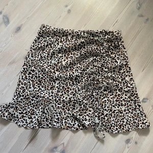 Leopardmönstrad kortkjol med volang - Snygg kortkjol med leopardmönster i beige, brunt och svart. Kjolen har volang nertill och en trendig snörning framtill som ger en cool detalj. Perfekt för dig som vill sticka ut med ett djurmönstrat plagg och gillar att leka med olika stilar.