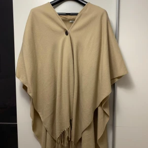 Beige poncho med fransar - Stilren beige poncho med en enkel knapp framtill och snygga fransar längs nederkanten. Perfekt att slänga över axlarna för en chill och trendig look. Tidlös design som passar till det mesta och är superenkel att matcha.