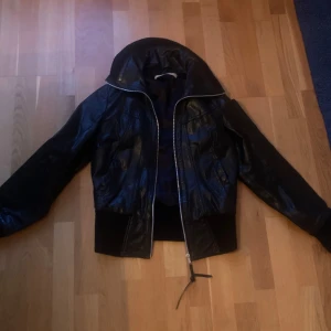 Svart skinnjacka Vero Moda - Säljer en svart bomberjacka från Vero Moda i skinnimitation med breda muddar vid midja och ärmslut. Jackan har dragkedja framtill och en hög krage som ger en cool vibe. Perfekt för dig som gillar streetstyle och vill ha en jacka med attityd.