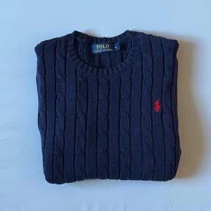 Mörkblå stickad tröja från Polo Ralph Lauren kabelstickad - Snygg mörkblå stickad tröja från Polo Ralph Lauren i storlek S. Tröjan har klassiskt kabelstickat mönster och en röd broderad logga på bröstet. Rund hals och långa ärmar, perfekt för dig som gillar stilrena och tidlösa plagg.
