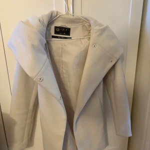 Beige kappa med stor krage från Amisu - Säljer en beige kappa från Amisu med stor, snygg krage och bälte i midjan. Jackan har långa ärmar och knappar framtill. Perfekt för dig som gillar stilrena och trendiga ytterplagg med lite extra attityd.